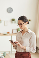 Vertical waist up portrait of elegant businesswoman using smartphone while standing in cozy home...