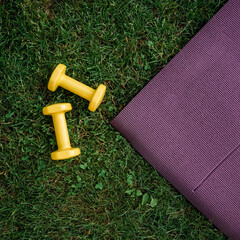 Set of two  yellow dumbbells and purple rubber Mat on a green grass background, top view. Staying in shape. Fitness and shape. Female exercise gear. Сoncept of outdoor fitness, nobody. Digital detox.