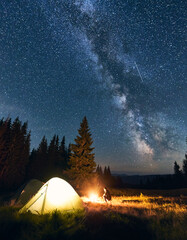 Lonely guy sitting near campfire in campsite under starry sky in the mountains. Night sky is strewn with bright stars and prominent Milky way. Tourist tents in valley with large pine trees.