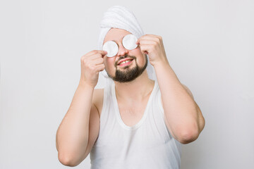 Happy smiling crazy funny bearded man with white towel oh his head, holding two cotton pads and hiding his eyes while caring about his skin. Close up portrait on white background. Spa for men