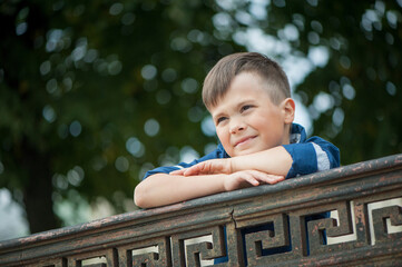 A handsome cute little boy in a blue shirt is leaning on an iron railing and looking into the distance, shot from below.