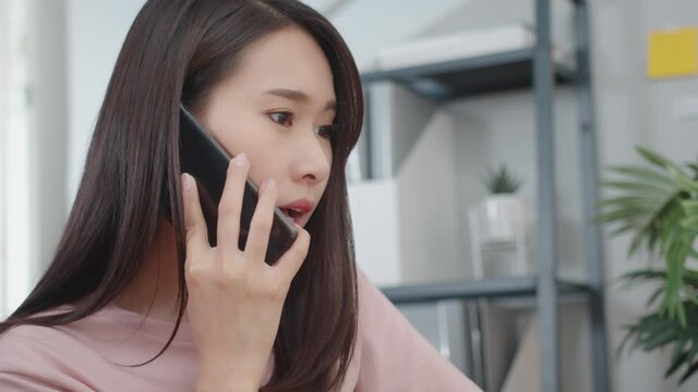 Attractive Asian Businesswoman talking on mobile phone with client sit at home office desk. Female Freelancer in casual clothes communicating with colleagues discussing a project work from home