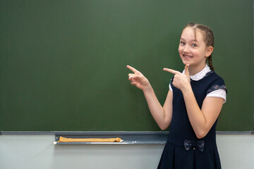 Back to school concept. Happy schoolgirl with backpack at the black chalkboard in classroom. Shows hands on copy space.