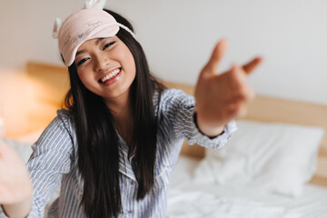 Joyful girl in blue pajamas and sleep mask poses in bedroom. Woman with smile reaches for camera