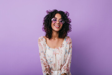 Indoor portrait of blithesome tanned woman with flowers in hair. Studio shot of glad african female model isolated on purple background.
