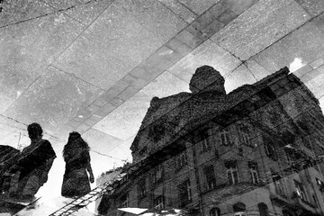 Blurry reflection shadow silhouettes in a puddle of a neoclassical building and two people walking on wet pedestrian street