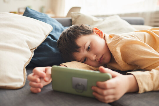 Warm-toned portrait of bored teenage boy using smartphone while lying on bed or couch at home, copy space