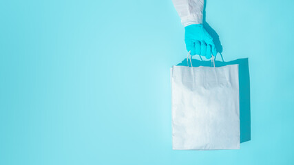 hand in gloves holding shopping paper bag. Concept of consumerism, shopping symbol during pandemic time. Copy space trend pastel color. Hard shadows, morning light.