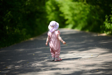A little girl in a hat and summer dress walks alone on the road, around the forest
