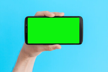 Smartphone with a green screen in left hand on an light blue background, horizontal position.