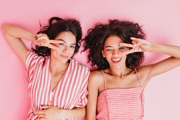 Beautiful and charming girls in cute pink pajamas lie on their backs in studio on gently pink background and show two fingers, in support of world peace
