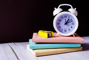 On a light table are diaries of pastel colors, on which stands a white alarm clock and a stapler