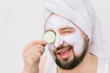 Close up portrait of unshaven bearded man in white towel oh his head, having cosmetic mask after shower, posing with slice of cucumber covering his eye, smiling and winking to camera