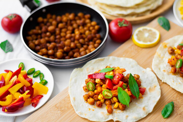 Vegan tortilla tacos with chickpeas, bell peppers, corn, tomatoes, basil leaves and green chilli. On the background are ingredients, lemons, chickpeas in a small pan