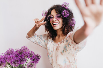 Enthusiastic african girl in stylish dress making selfie with alliums. Spectacular black young woman having fun during indoor photoshoot with purple flowers on white background.