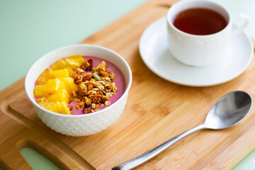 Healthy summer breakfast of a pink smoothie bowl made with frozen banana and raspberry garnished with granola and fresh orange. In the background is black tea