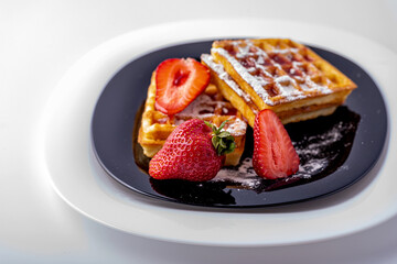Belgian waffles with strawberries and mint on a black plate and white background.