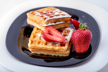Belgian waffles with strawberries and mint on a black plate and white background.