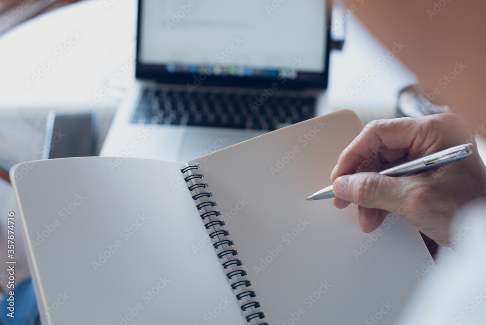 Wall mural Mockup of man with pen in hand writing on blank page notebook while working on laptop computer at coffee shop