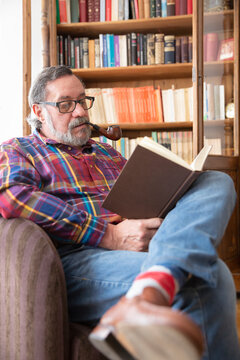 Portrait of older man reading a book next to his own library while smoking a pipe. Stay home concept.