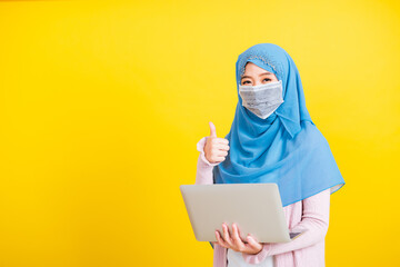 Asian Muslim Arab, Portrait of happy beautiful young woman Islam religious wear veil hijab and face mask protect she quarantines coronavirus hold laptop computer and show finger thumb up for good sign