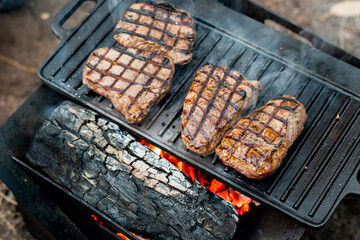 Beef steaks grilling on a cast iron plate on a camp fire. Campfire cooking. Outdoor BBQ. Selective focus.