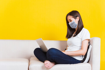 Fototapeta na wymiar Portrait Asian of beautiful young woman sitting on sofa wearing medical face mask protective she work from home with laptop computer during Coronavirus studio shot isolated on yellow background