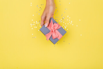 Women hands holding a gift or gift box decorated with confetti on a yellow background top view.