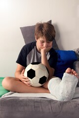 Bored boy with a broken leg sitting on the couch and holding a football ball.A boy with a broken...