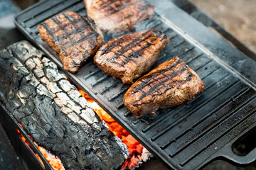 Beef steaks grilling on a cast iron plate on a camp fire. Campfire cooking. Outdoor BBQ. Selective focus.