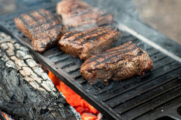 Beef steaks grilling on a cast iron plate on a camp fire. Campfire cooking. Outdoor BBQ. Selective focus.