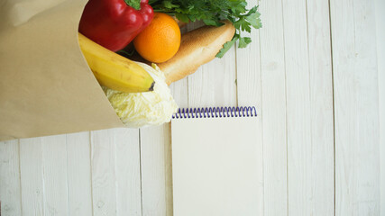 Full paper bag of food products with blank little notebook on wooden table, top view