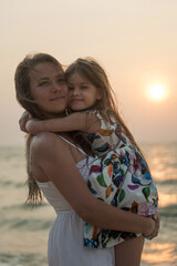 Fototapeta na wymiar Smiling mother and beautiful daughter having romantic evening on the beach during sunset. Portrait of happy woman hugging to cute little girl on the sea. Portrait of happy blonde kid embracing her mom