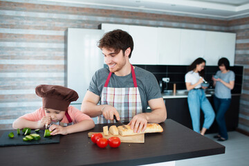 Happy family is cooking in the kitchen. Father teaches daughter to cut vegetables. Mother and grandmother stand in the background