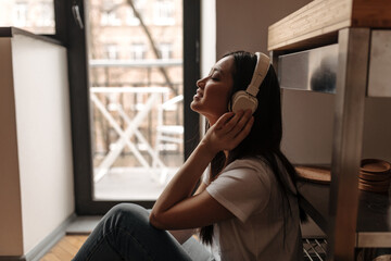 Lady in white massive headphones listens to song while sitting on floor in apartment against window