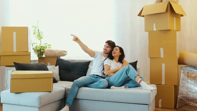 Young couple move into new apartment. Beautiful couple sit together on sofa and having fun. Guy pointing forward. Camera moves to the back. Making plans for apatment and furniture.