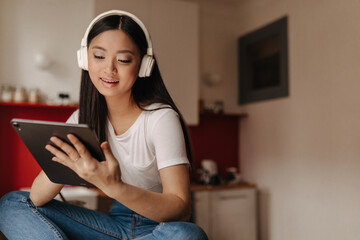 Obraz premium Lady in massive headphones with sweet smile looks into tablet while sitting in kitchen