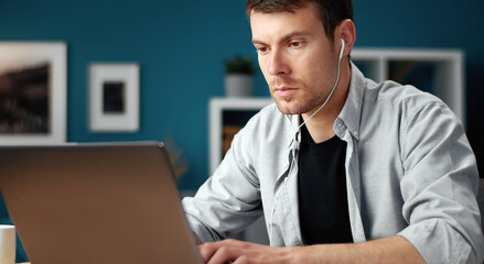 Portrait of focused young man with earphones using laptop sitting at desk at home, half-turn shot