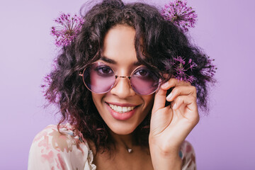 Close-up portrait of smiling brown-eyed woman with flowers in black hair. African blissful lady in sunglasses posing on purple background.