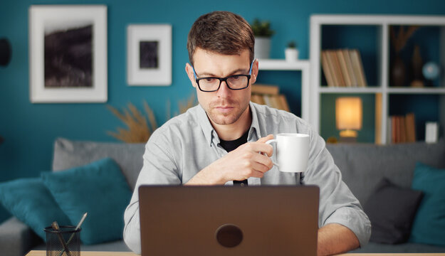 Front View Of Involved Young Man Browsing Laptop And Holding Cup Sitting At Table In Living Room