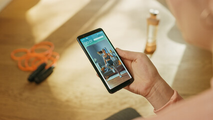 Over Shoulder Shot: Fit Woman Holds Smartphone, Uses Fitness Tracking App, Looking Premium Video Explaining Exercise. Mock-up Training Application Software