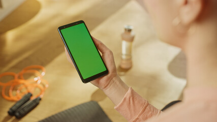 Fitness Concept: Woman Holding Chroma Key Green Screen Smartphone. In the Background Fitness Mat, Jumping Rope and Equipment. Close-up Over The Shoulde Shot.