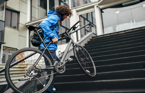 Rear view image of young man with curly hair climbing on the building's stairs with his bike. Male courier with curly hair delivers parcel with a bicycle on the city.
