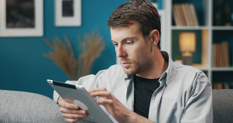 Half turn portrait of young casual man using tablet touching screen sitting on sofa