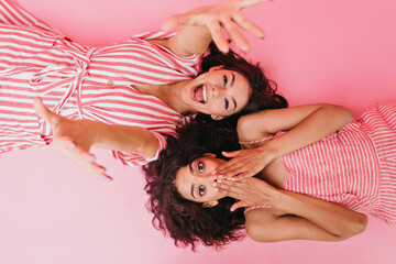 Obraz na płótnie Canvas Girlfriends with dark curly hair are having fun at their pajama party. Laughing girl stretches towards camera while her cousin covers her face in surprise