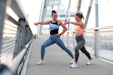 Fototapeta na wymiar Two girl exercise with weights on hands at bridge at the end of day on sunset