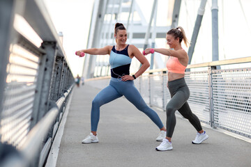 Two girl exercise with weights on hands at bridge at the end of day on sunset