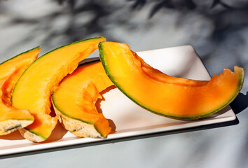 Honey melon or cantaloupe (Cucumis melo). Sliced of Japanese melon on white plate with shadows. Summertime fruits. Healthcare concept. Selective focus