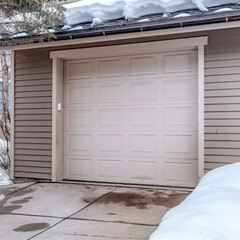 Square frame White garage door entrance of home with snow covered roof and yard in winter