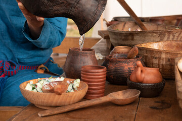 The girl pours water from the jug into a clay mug on a wooden table. Historic folk dishes and natural food.